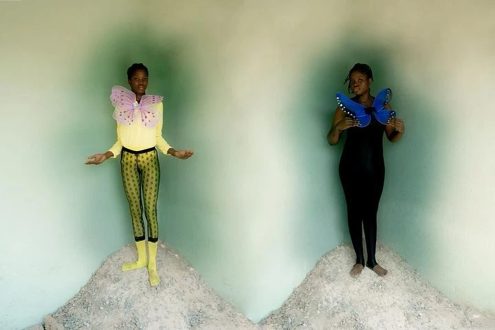 Two women stand on small piles of gravel against an ombre green wall, each wearing colorful butterfly wings and full body suits.