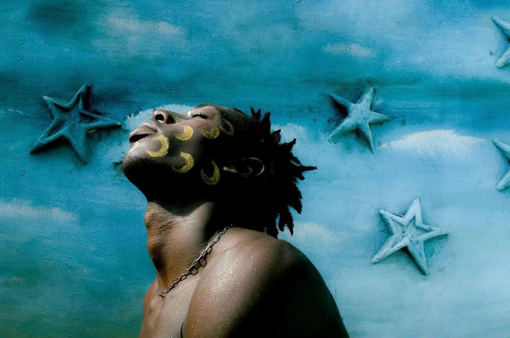 Portrait of African-descent adult male with yellow face paint and chain necklace, looking up against a blue star-studded backdrop.