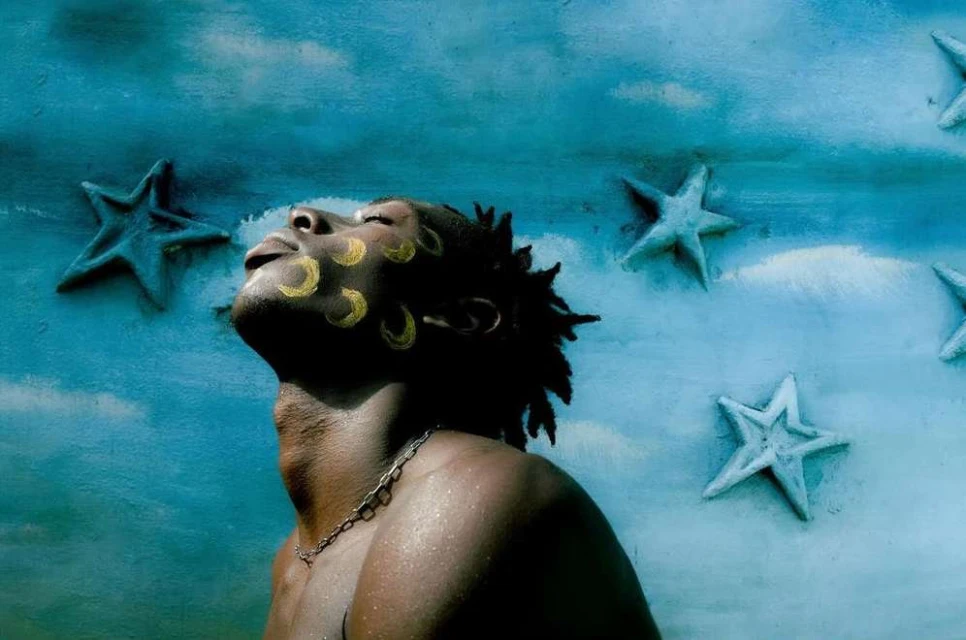 Portrait of African-descent adult male with yellow face paint and chain necklace, looking up against a blue star-studded backdrop.