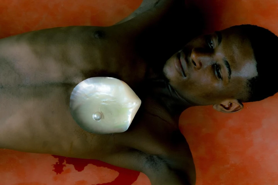 A young adult with an expressionless face lays against an orange backdrop with a large pearlescent shell over his chest.
