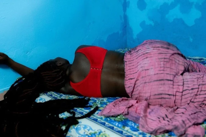 Black woman with long dark dreadlocks lying on a bed wearing a red bra and pink patterned trousers against a blue wall