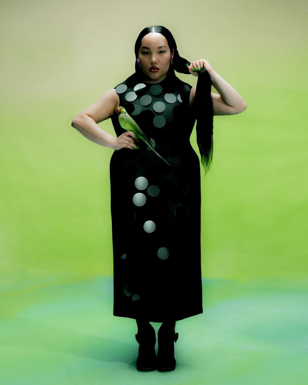 An Asian woman poses in a black dress with white polka dots, holding a green parrot in a studio with a blended yellow-green backdrop.