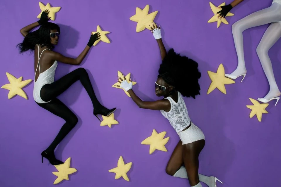 Three fashion models on purple background holding large yellow star props wearing leotards, tights, gloves, and sunglasses.
