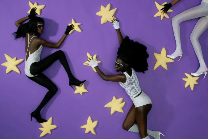 Three fashion models on purple background holding large yellow star props wearing leotards, tights, gloves, and sunglasses.