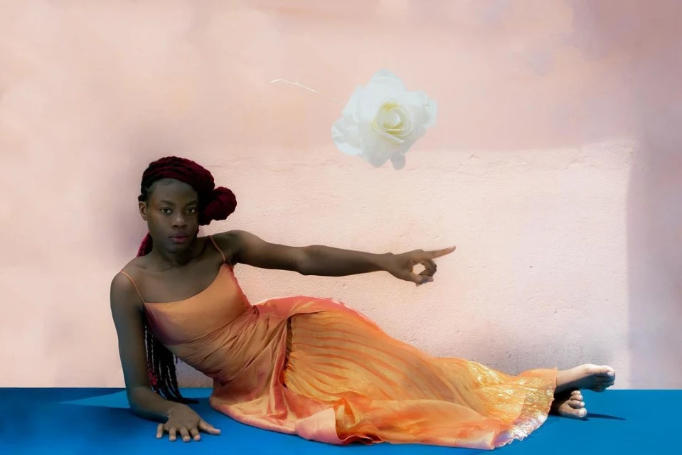Elegant woman in a dress points at a large floating white flower against a pink and white backdrop, creating a surreal, artistic scene.