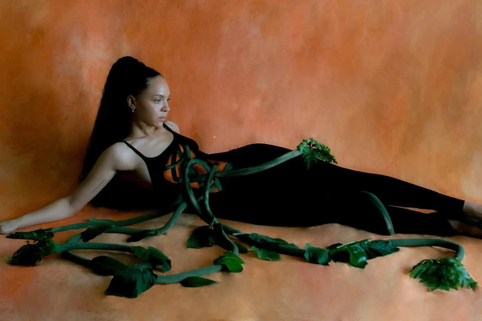 Adult woman in black bodysuit lying on an orange surface among leafy green vines in a fashion photography style pose.