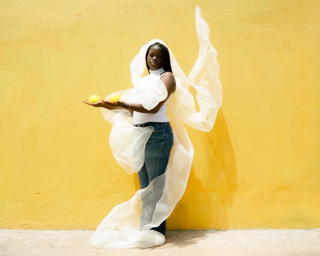 An African-American woman with a white wrap, wearing a white top and jeans, holds two lemons in front of a yellow wall in an outdoor setting.