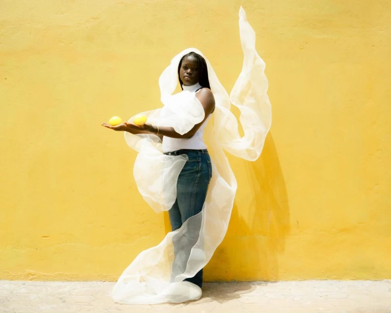 An African-American woman with a white wrap, wearing a white top and jeans, holds two lemons in front of a yellow wall in an outdoor setting.