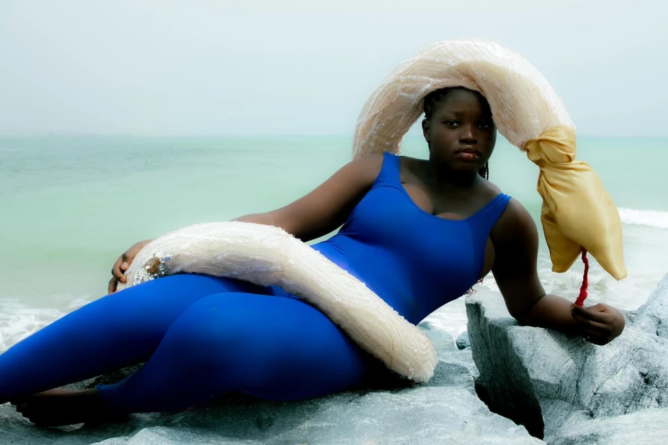 A Black woman in a blue bodysuit and a unique headpiece poses on a rocky beach with the ocean and sky creating a serene backdrop.