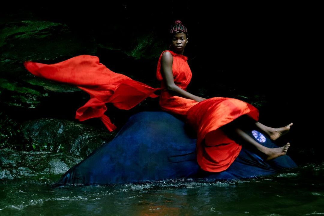 An elegant Black woman in a red dress sits on a rock in a stream. The fabric flows around her in a dark, natural environment.