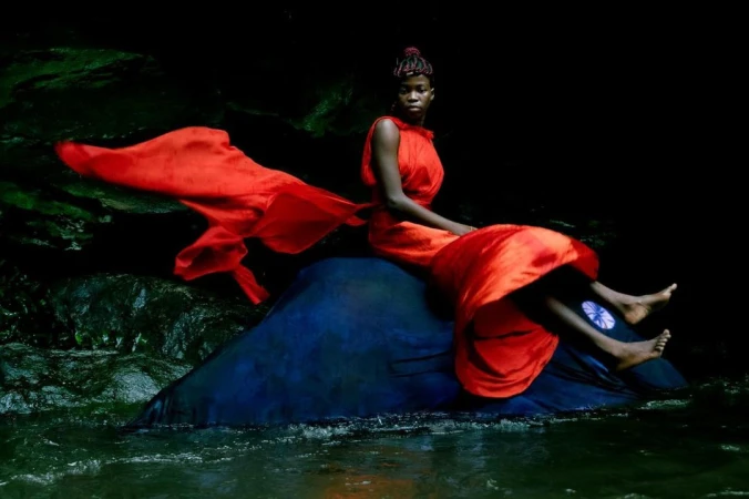 An elegant Black woman in a red dress sits on a rock in a stream. The fabric flows around her in a dark, natural environment.