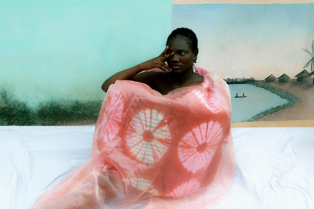Portrait of an African woman wrapped in a patterned textile, seated against a split blue and painted landscape background, in a studio setting.