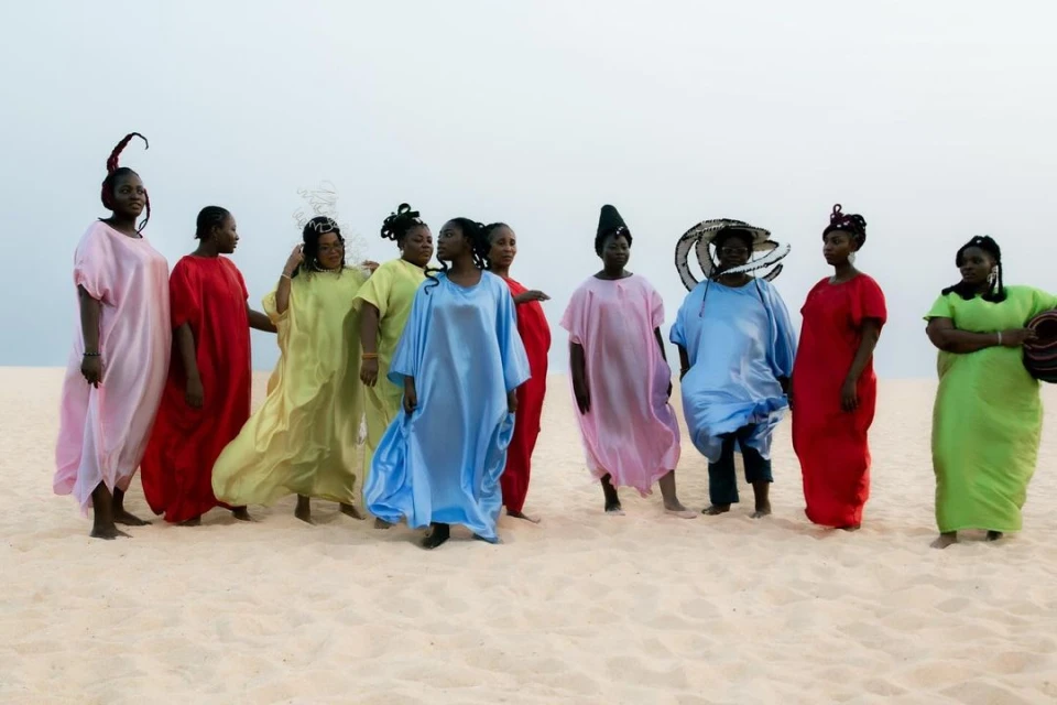 Group of ten women in colorful, oversized gowns on a sandy beach with a neutral sky backdrop, showcasing simplicity and modest fashion.