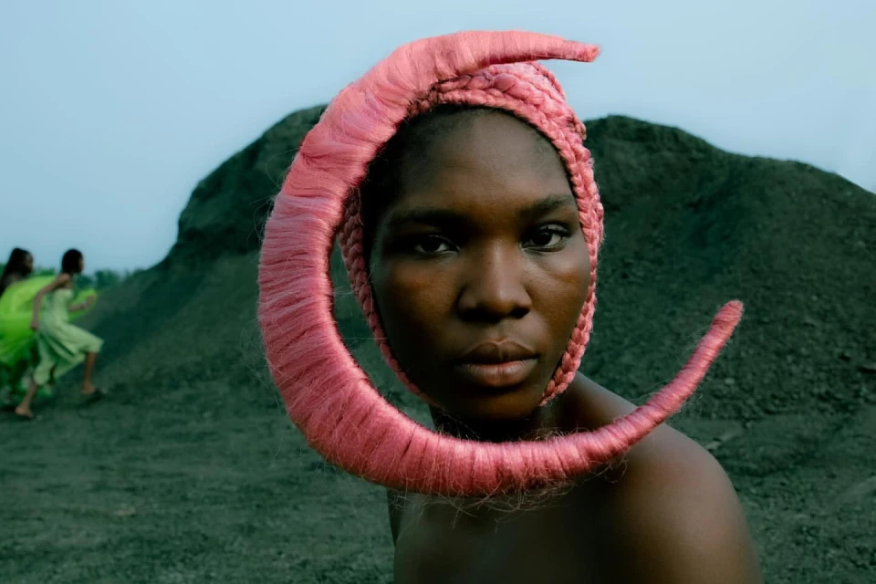 Woman with unique pink headdress poses in a portrait on a dark grassy plain with people in green dresses and mounds in background.