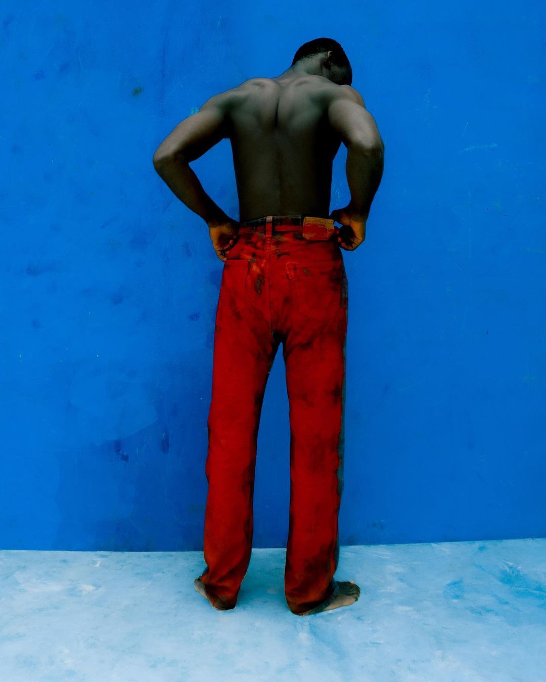 Back view of a shirtless Black man in red pants standing against a blue wall. His hands are on his hips in a studio shot.