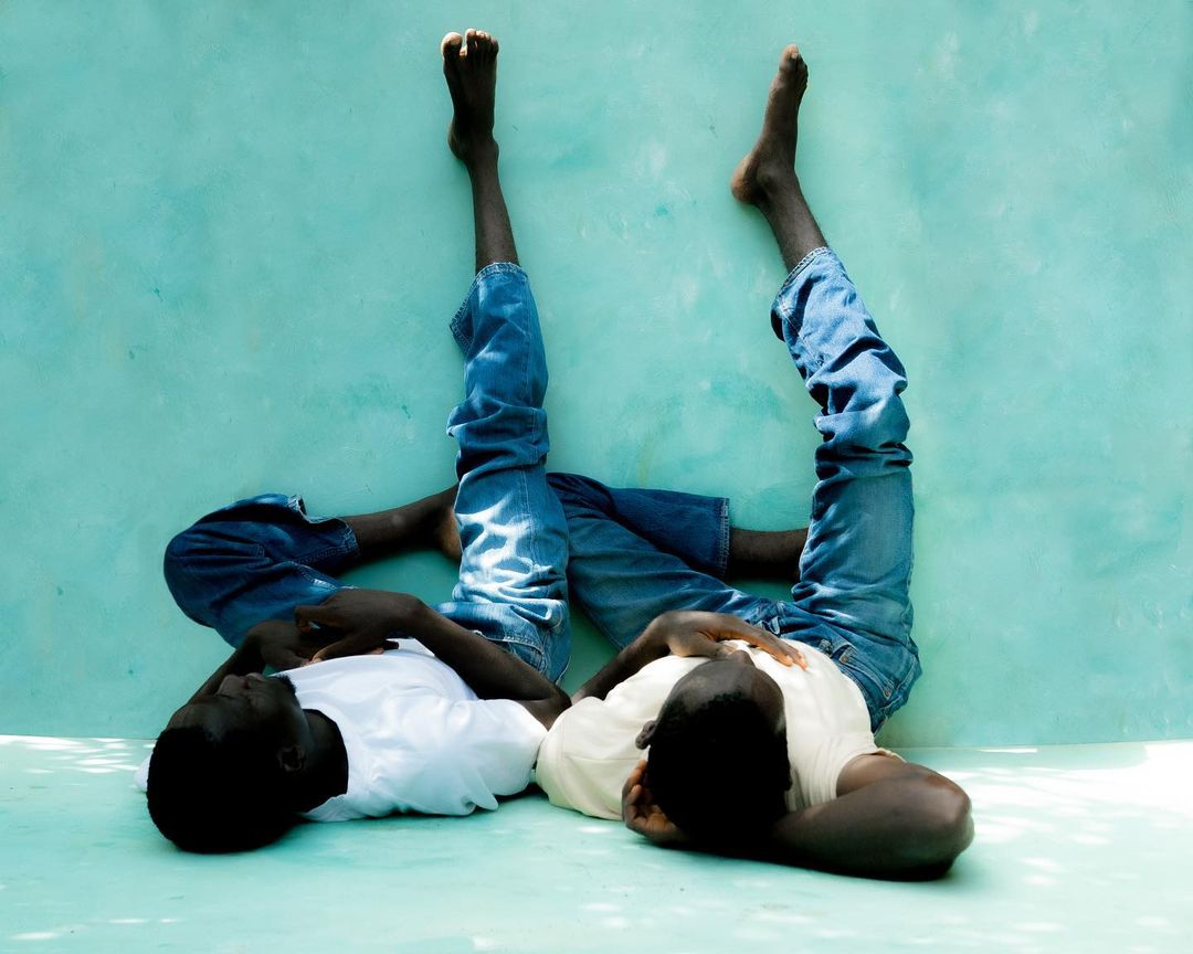 Two dark-skinned young adults lie on their sides against a blue wall. One person's bare feet extend upward as legs rest on the wall.