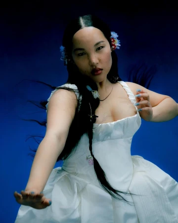 Woman in a white dress with dark hair and blue nails posing against a deep blue background by Delali Ayivi