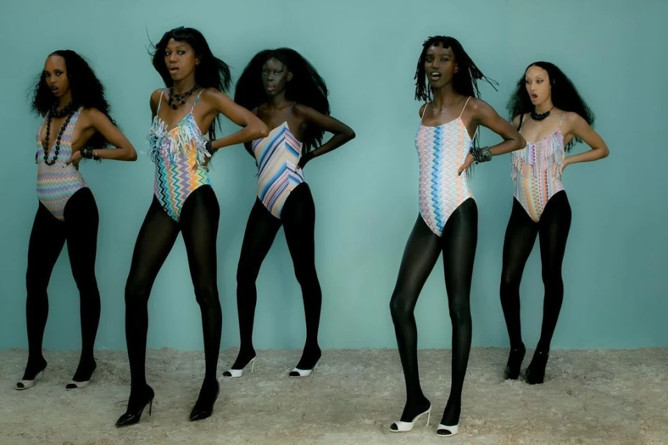 Five dark-skinned women in patterned bodysuits and black tights strike a pose against a blue backdrop. Fashion, style, and visual arts.