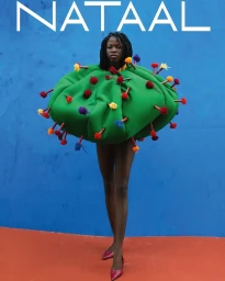 Model wearing a round green padded sculptural garment with colorful pom-pom pins against a blue wall