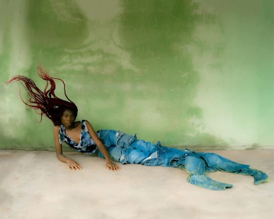 Black woman with red braided hair lying on floor against weathered green wall wearing blue denim patchwork pants