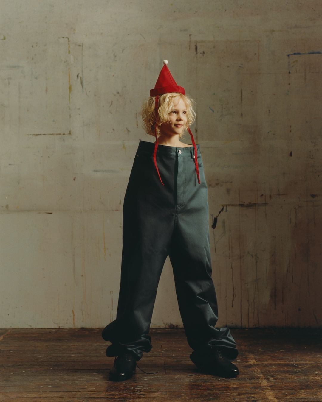 A child with blonde curly hair wearing a red hat stands in oversized pants and black shoes against a textured white wall and wood floor.
