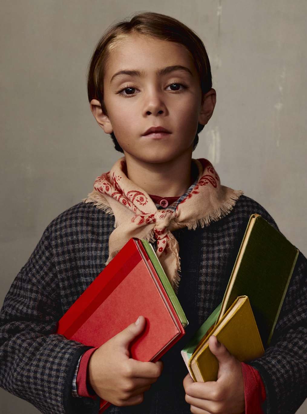 Portrait of a young boy with a scarf, dressed in a grey checkered coat, holding several books against a neutral-toned background.