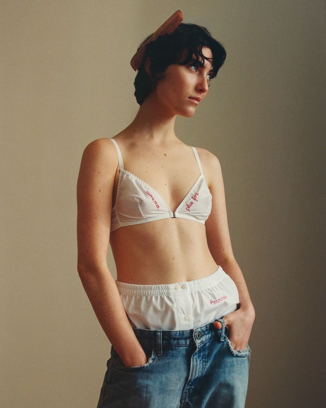 A young adult woman poses indoors, wearing a white bra and boxer shorts with the waistband visible above blue jeans. She has a comb in her hair.