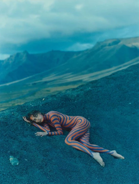 Woman with body paint lies on a hill with mountains in the background. Conceptual fashion and art photography.