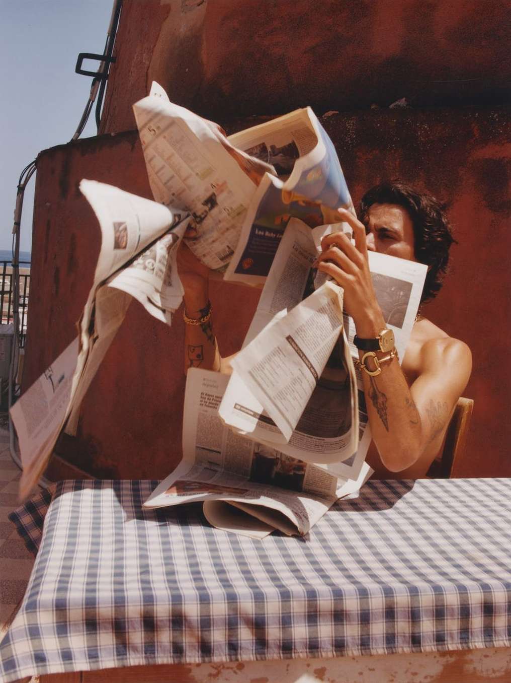 Adult male sits at a table with a blue-and-white tablecloth, surrounded by scattered newspapers, outside during the daytime.