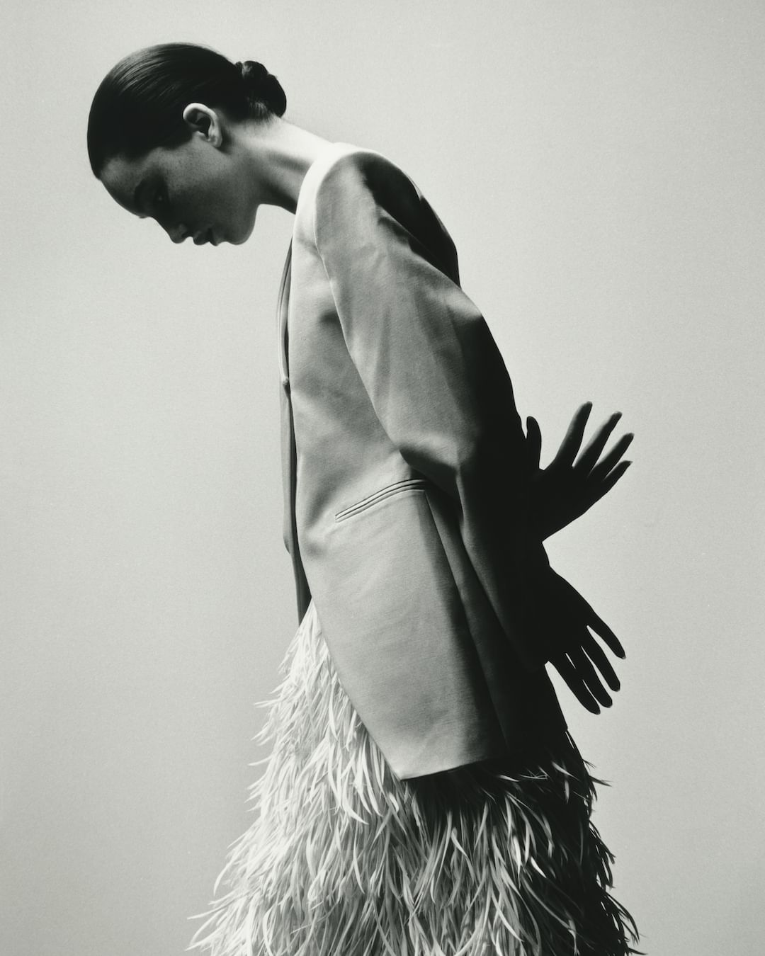 A monochrome fashion shot of a woman in a feathered skirt and blazer, standing against a neutral backdrop with hand shadow.