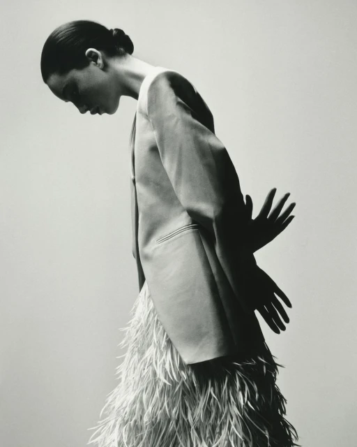 A monochrome fashion shot of a woman in a feathered skirt and blazer, standing against a neutral backdrop with hand shadow.