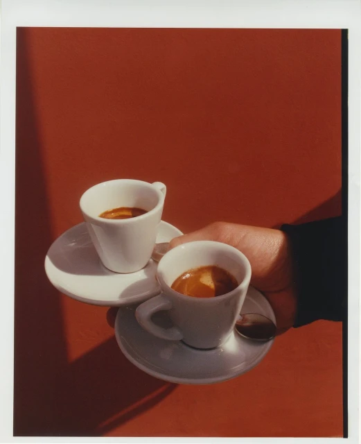 Close-up studio shot of two cups of espresso held by a hand. A spoon rests on one saucer against a solid red background.
