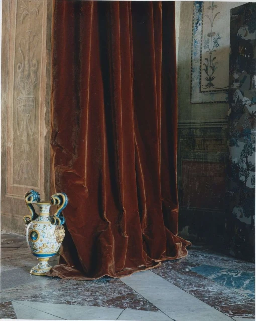 A decorative vase sits in an ornate interior with reddish-brown velvet curtains and decorative wall molding creating a rich, historical atmosphere.