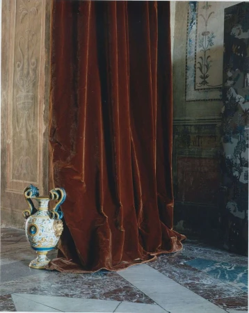 A decorative vase sits in an ornate interior with reddish-brown velvet curtains and decorative wall molding creating a rich, historical atmosphere.