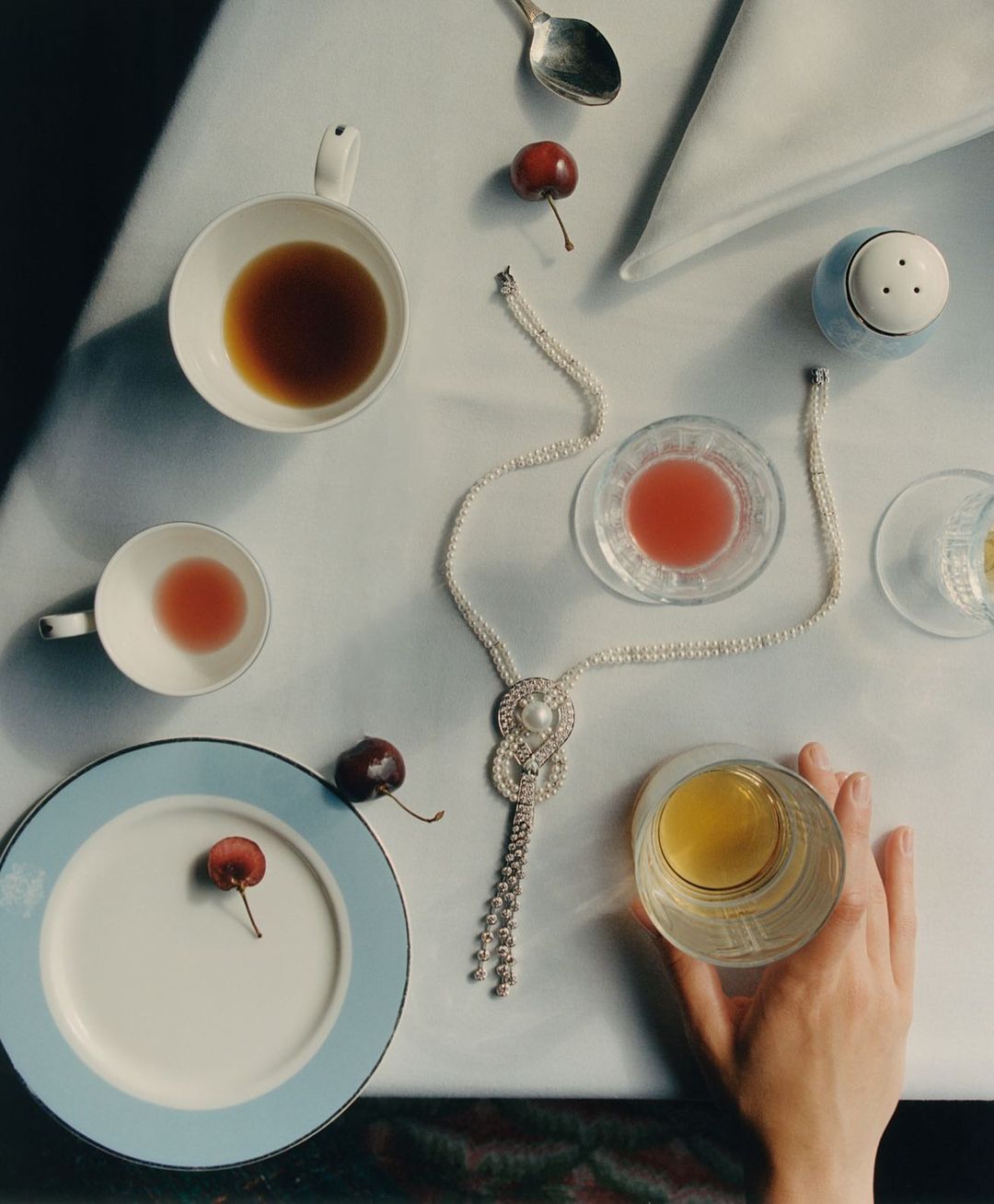 Still life on a table with a hand holding a glass of orange liquid, complemented by cherries, cups, plates, and a pearl necklace.