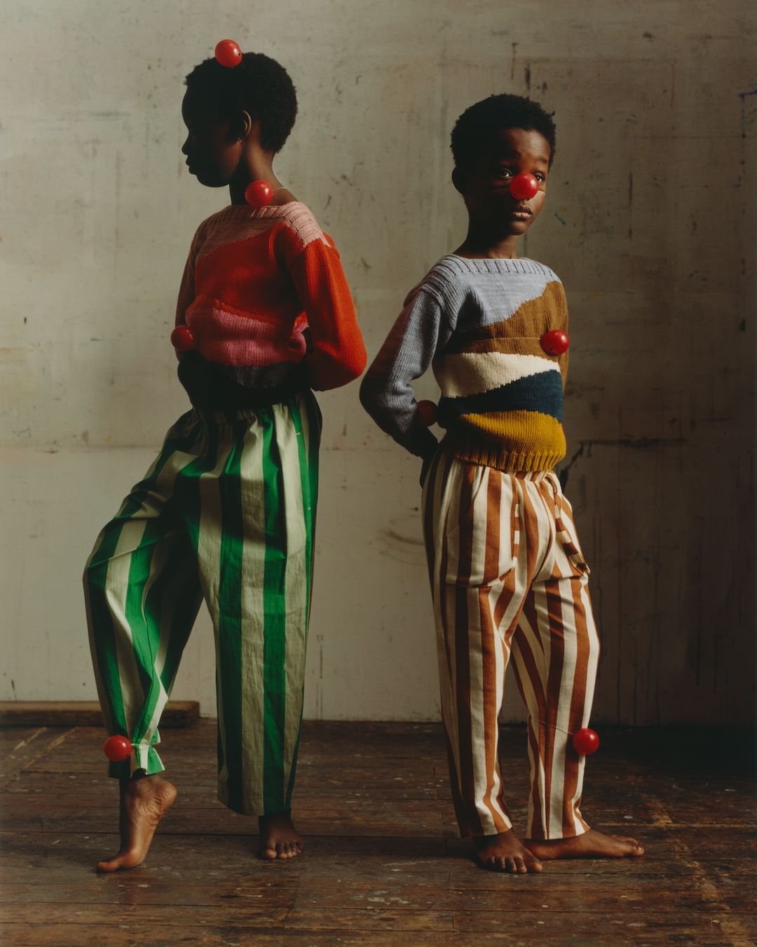 Two young children pose indoors wearing sweaters and striped pants with red balls attached to their bodies and faces, studio lighting.