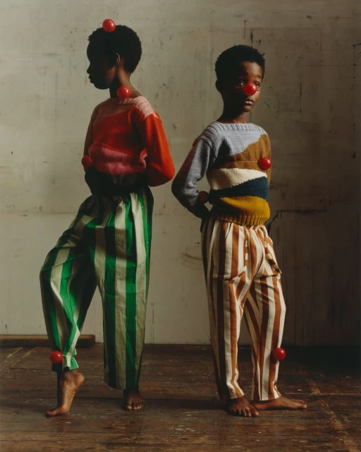 Two young children pose indoors wearing sweaters and striped pants with red balls attached to their bodies and faces, studio lighting.