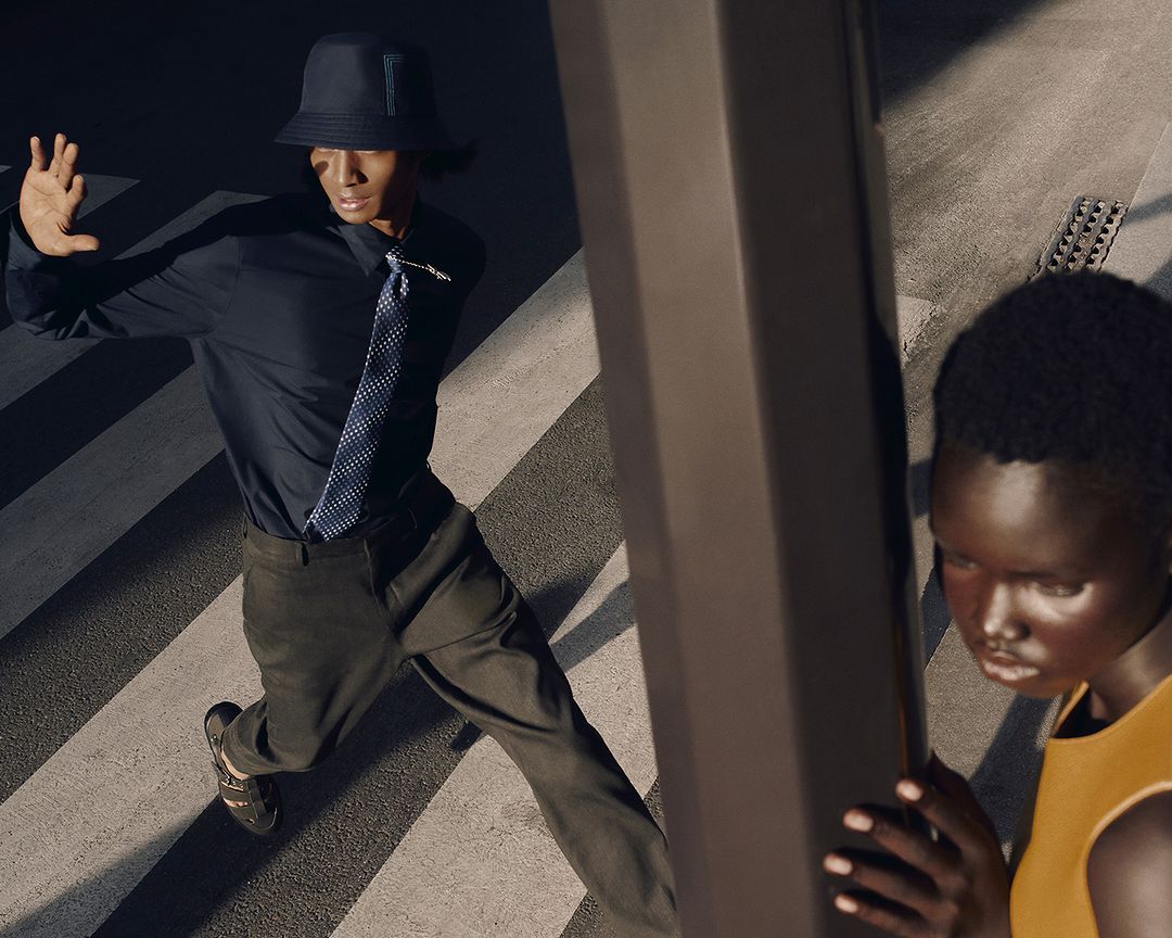 Two fashion models in stylish outfits pose on a crosswalk in an urban setting with a metal beam, creating a modern editorial shot.