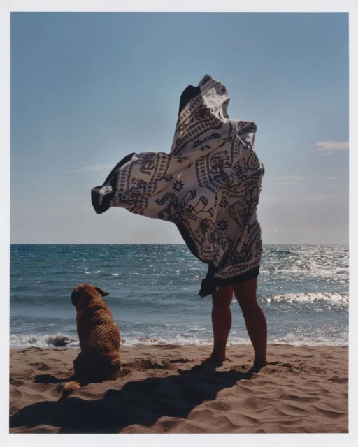 Dog sitting next to a person draped in patterned textile on a sandy beach with blue ocean and sky in the background on a sunny day.