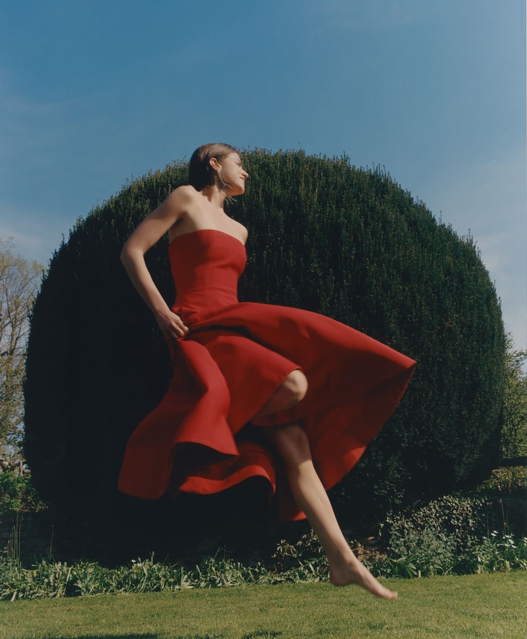 Young woman in red strapless dress twirling barefoot on grass in outdoor shot, large green bush and blue sky in background.