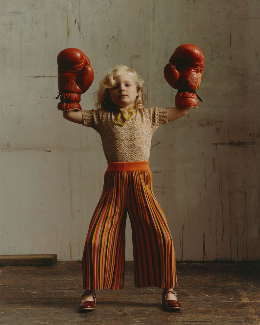 Blonde child in retro-style striped culottes and Mary Janes strikes a playful pose with oversized vintage boxing gloves indoors.