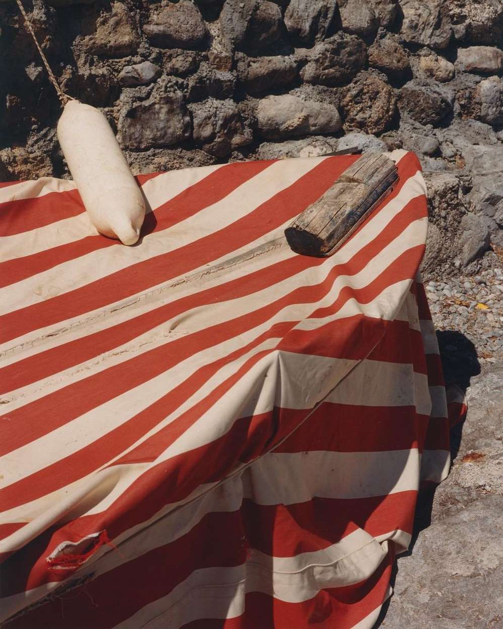 Still life of a boat cover with red and white stripes, a white boat fender, and a piece of wood, set against a stone wall and ground.
