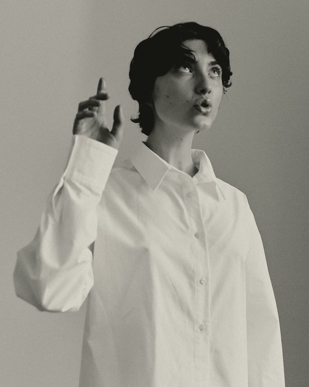 Monochrome portrait of a young adult woman with short dark hair in a white collared shirt, looking up and pointing with her left hand.