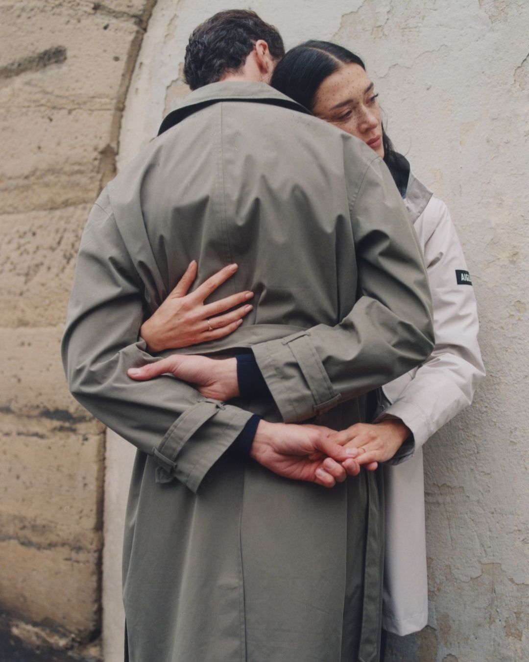 A couple embraces on a city street, wearing trench coats in muted tones. The woman looks downward while the man's back is to the camera.