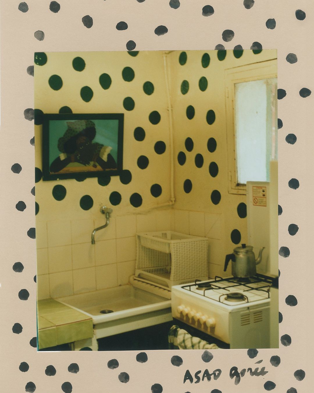 Interior view of a kitchen with green polka-dot wallpaper, a vintage stove, metal sink, window, and a framed picture on the wall.