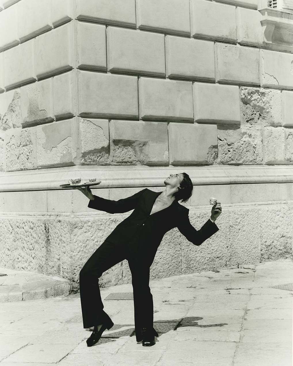 Elegant server in black suit balances tray and glass against a stone building and tiled pavement in a monochrome photograph.