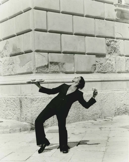 Elegant server in black suit balances tray and glass against a stone building and tiled pavement in a monochrome photograph.