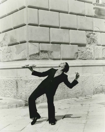 Elegant server in black suit balances tray and glass against a stone building and tiled pavement in a monochrome photograph.