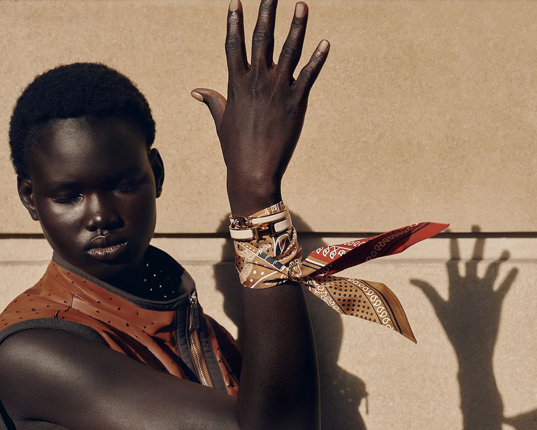 Close-up shot of a Black female model with a patterned silk scarf tied around her wrist, posing against a neutral wall with hand shadow.