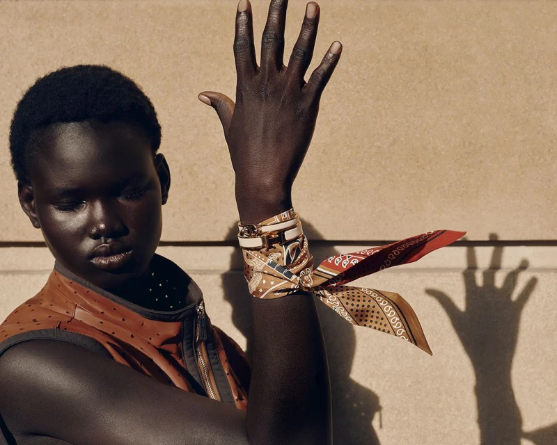 Close-up shot of a Black female model with a patterned silk scarf tied around her wrist, posing against a neutral wall with hand shadow.